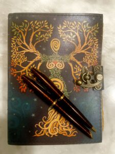 A leather journal with an abstract illustrated tree on the front with two pens laying on top.